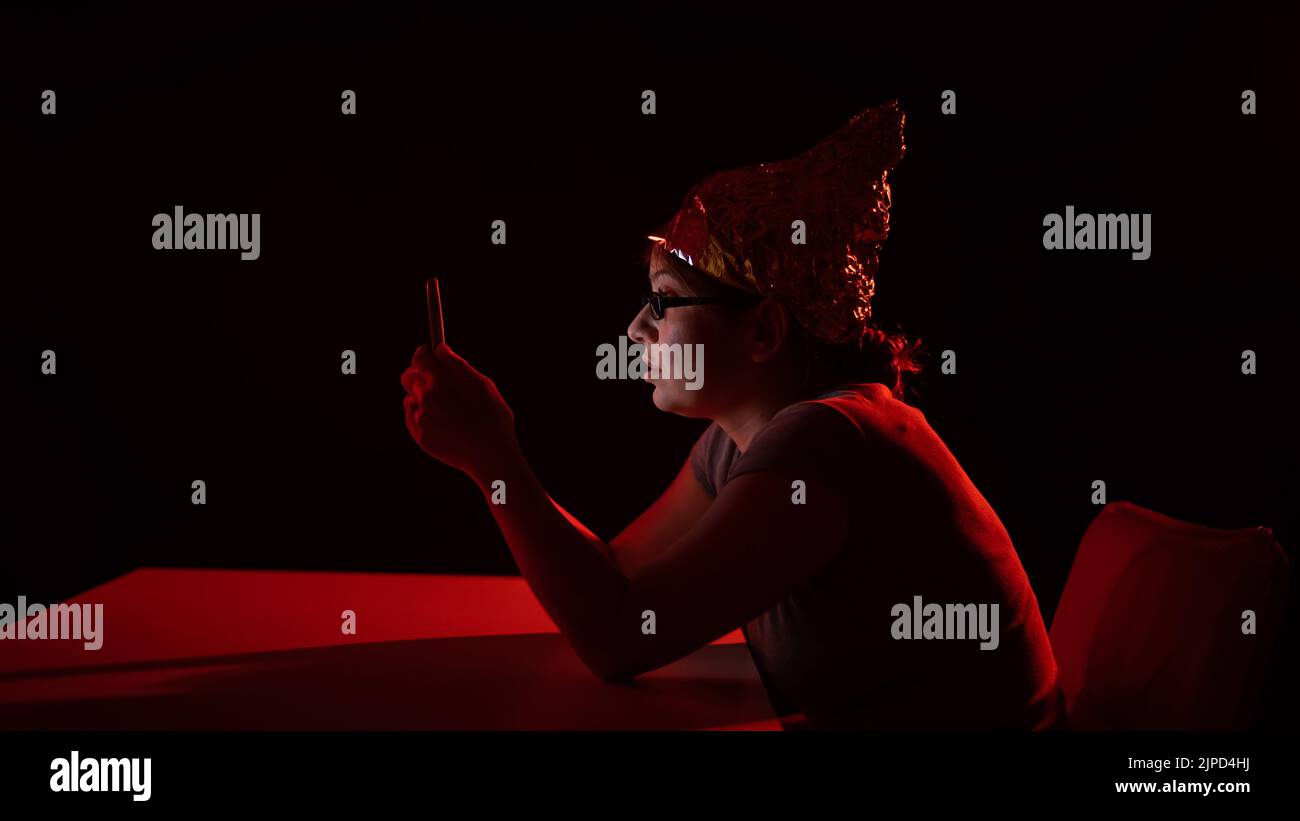 Paranoid caucasian woman using smartphone wearing tinfoil hat. Conspiracy theory.  Stock Photo