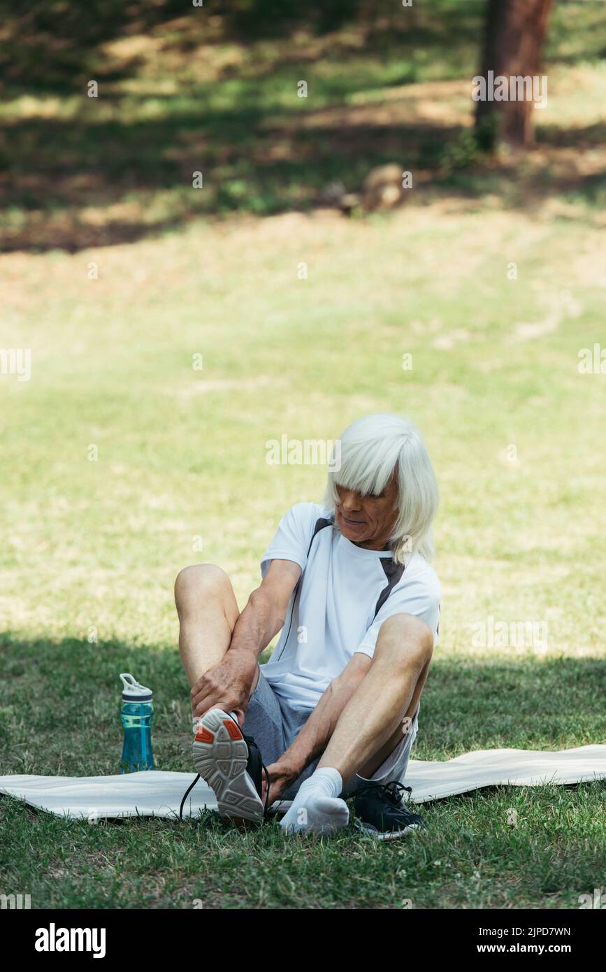 senior man with grey hair taking off sneakers while sitting on fitness mat in park Stock Photo