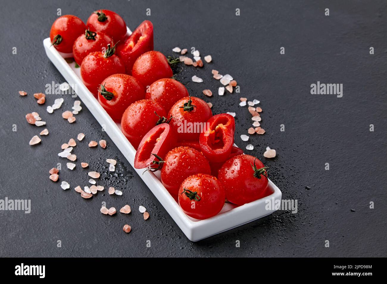 An elongated dish of tomatoes and paprika with drops of water or dew and coarse pink Tibetan salt on a black concrete table. High angle with copy spac Stock Photo