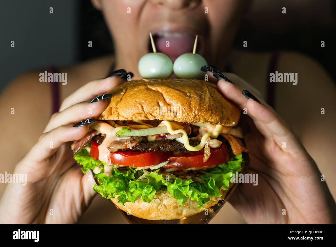 latina girl about to bite a hamburger she is holding with both hands. hamburger with two quail eggs on top imitating eyes. street food on black backgr Stock Photo