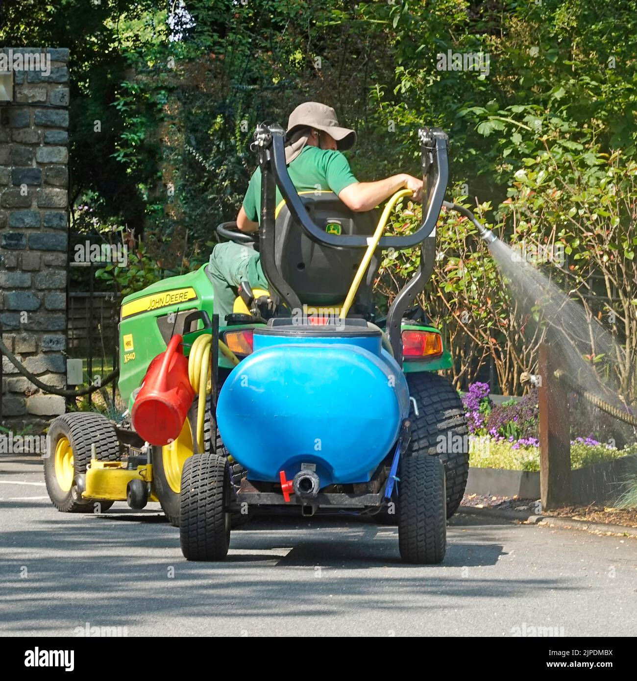 Gardener sitting high up on John Deere mini tractor towing a blue mini pumped water tanker trailer spraying flowers in hotel country estate England UK Stock Photo