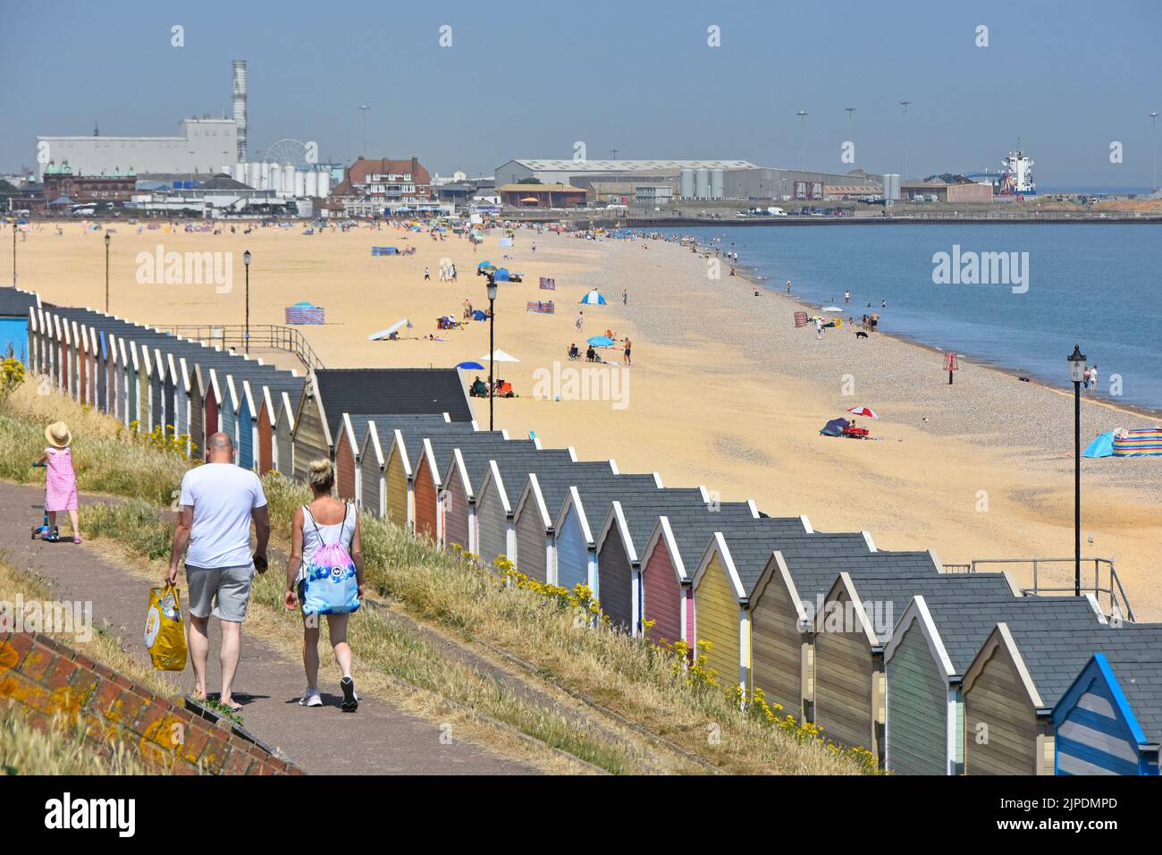 Couple & young girl on scooter walk cliff top path at Gorleston-on-Sea beach hut resort view towards Great Yarmouth industrial landscape Norfolk UK Stock Photo