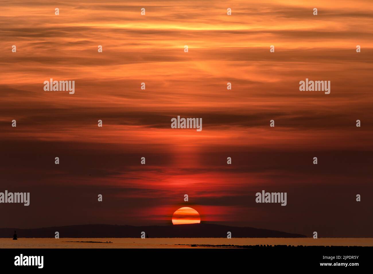 A setting sun behind a hill with the sea in the foreground.  The sky is various shades of red and orange. Stock Photo