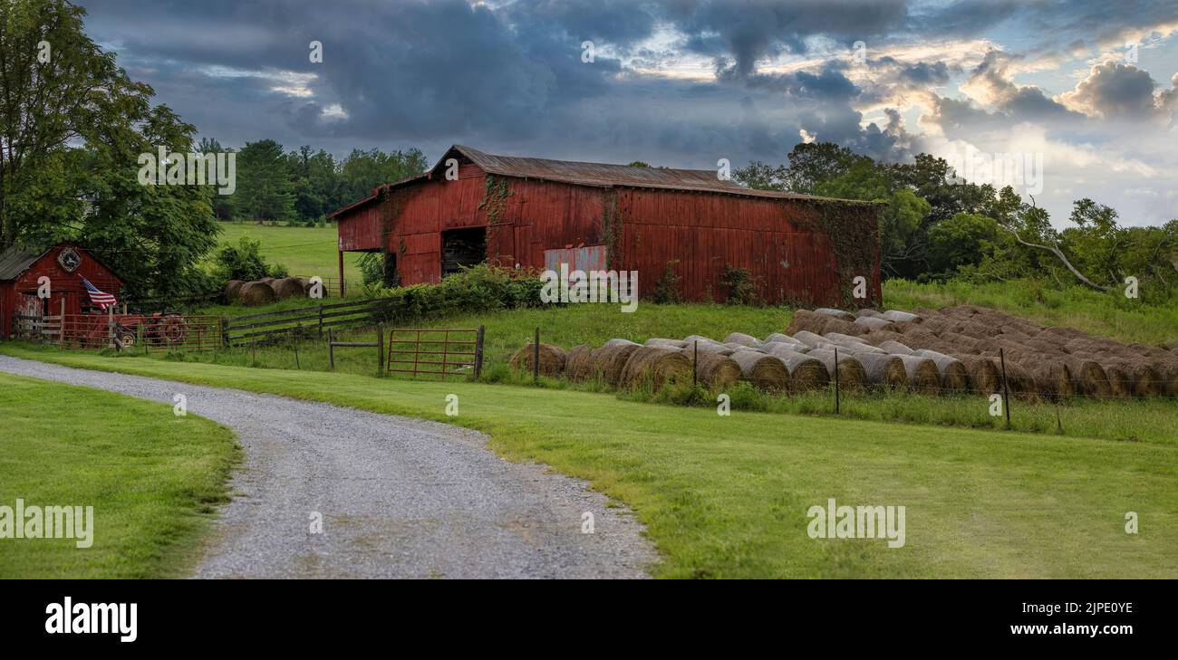 Seen from a country rural road a gravel road leading to a old red barn and out building with American Flag and rolled grass bails. Stock Photo