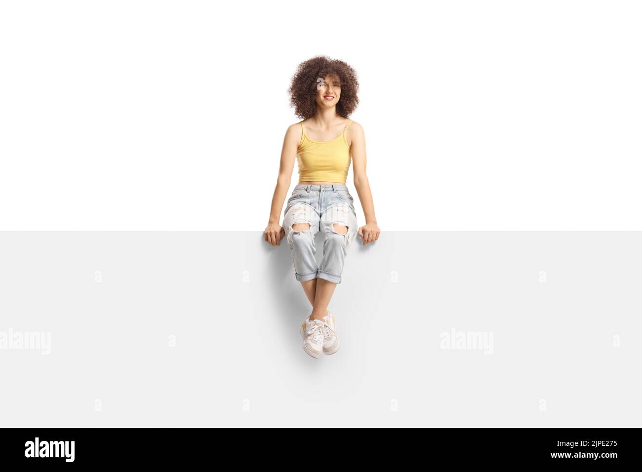 Full length portrait of a trendy young Caucasian female with afro hairstyle sitting on a blank panel isolated on white background Stock Photo