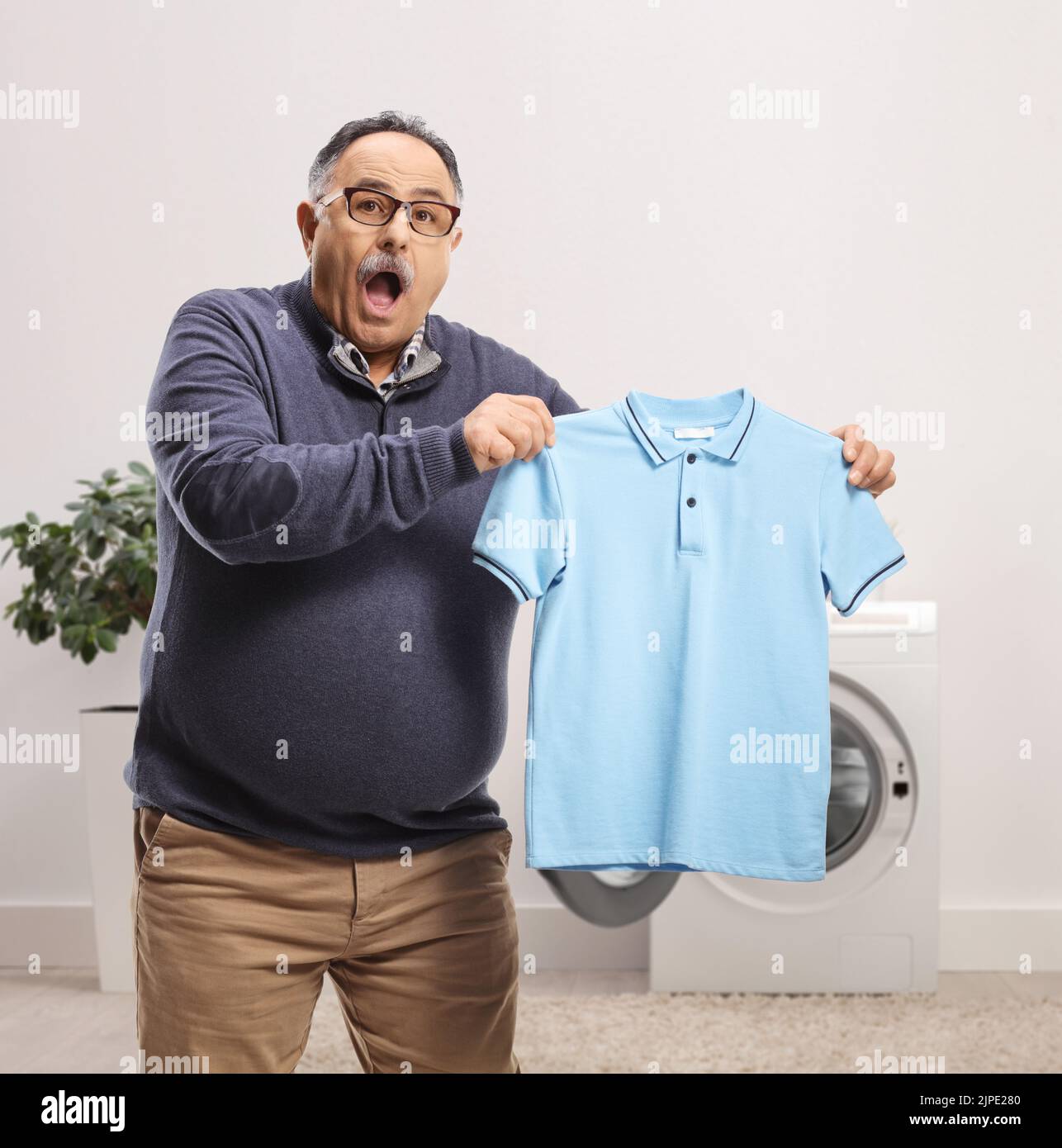 Confused mature man holding a shrunken t-shirt in front of a washing machine Stock Photo
