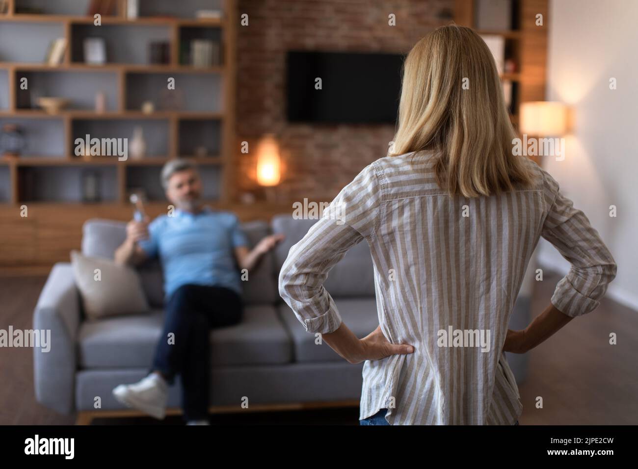 Unhappy sad adult caucasian wife angry at drunk husband with bottle in living room interior, back, blurred Stock Photo