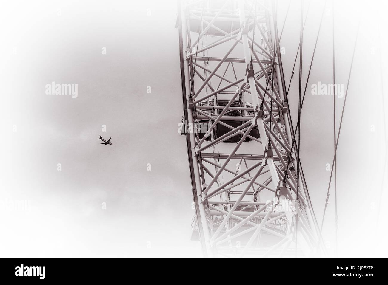 A monochrome image of a plane flying in the sky with part of the London Eye in the foreground. Stock Photo