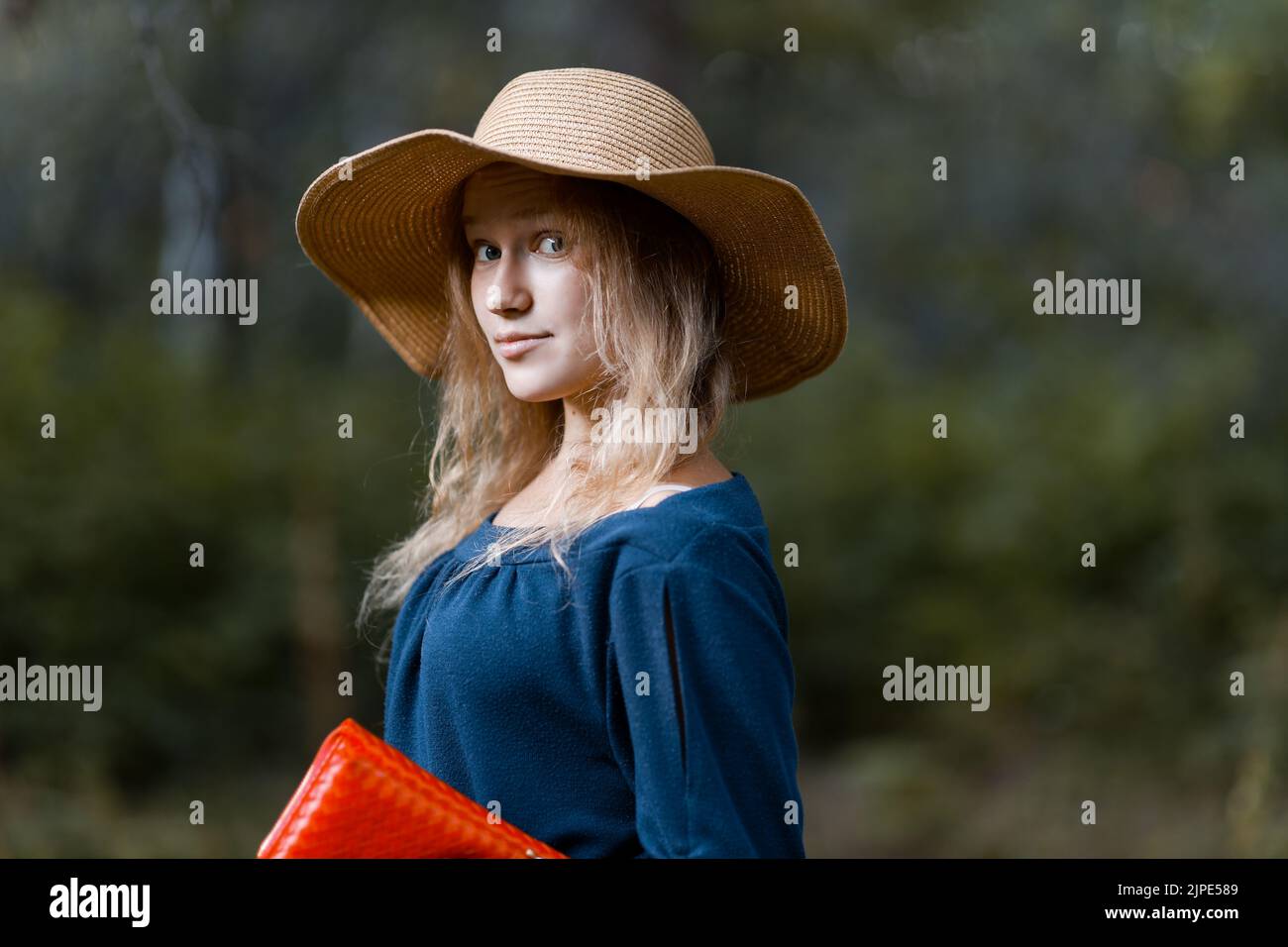 Closeup portrait of a young longhair girl in a straw hat and with a red bag in her hand against blurred nature background. Copyspace. Stock Photo