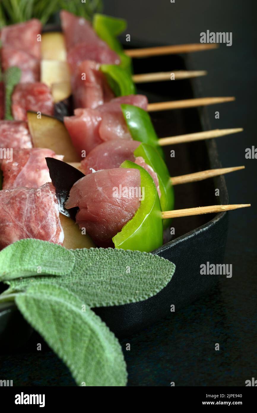 Macro close up of raw meat skewers before cooking on a dark background with sage leaves and mixed pepper. Stock Photo