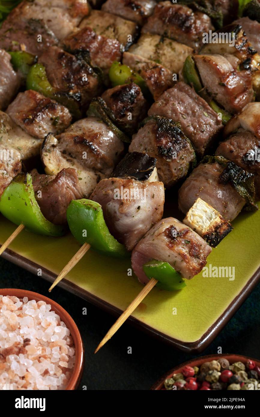 Macro close up of baked meat skewers on a green dish with sage leaves, salt and mixed pepper. Stock Photo