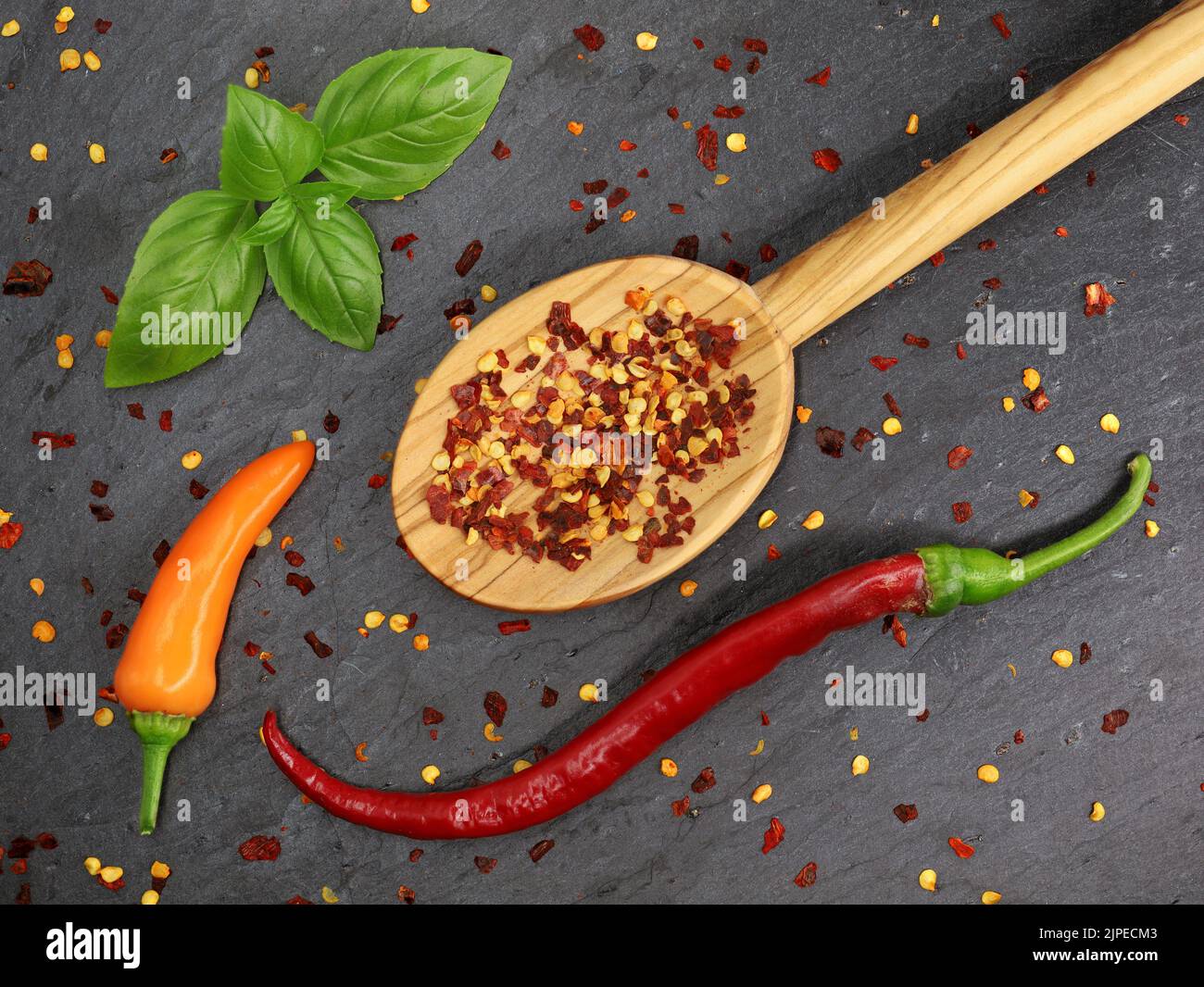 Top view of dried chili flakes and seeds in wooden cooking spoon with red and orange chillies on black slate board Stock Photo