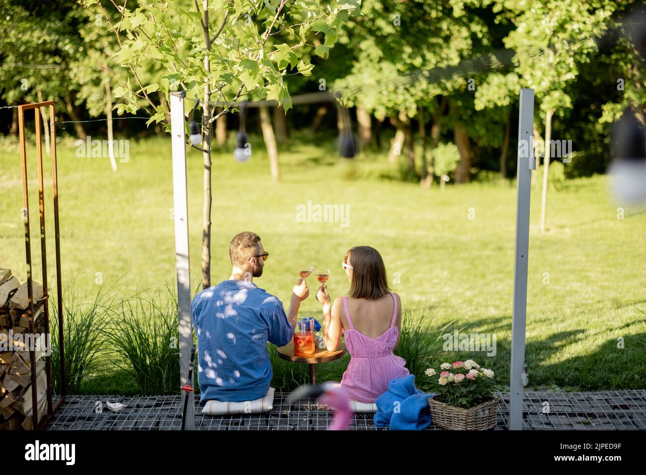 Young couple sit on porch of country house, have a drink and enjoy beautiful view on nature, view from the backside. Man and woman spend summer time r Stock Photo