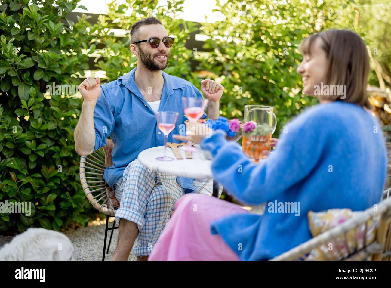 Young stylish couple have conversation while sitting by the round coffee table at their beautiful garden. Young man and woman have romantic evening ou Stock Photo