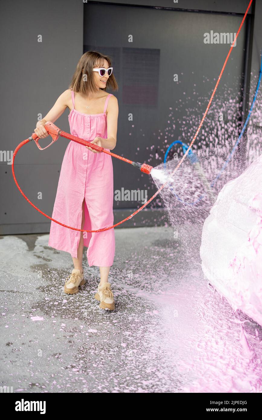 Young woman in pink dress washing her tiny car with nano foam at car wash during summer time. Easy and beautiful self-service at car wash concept Stock Photo