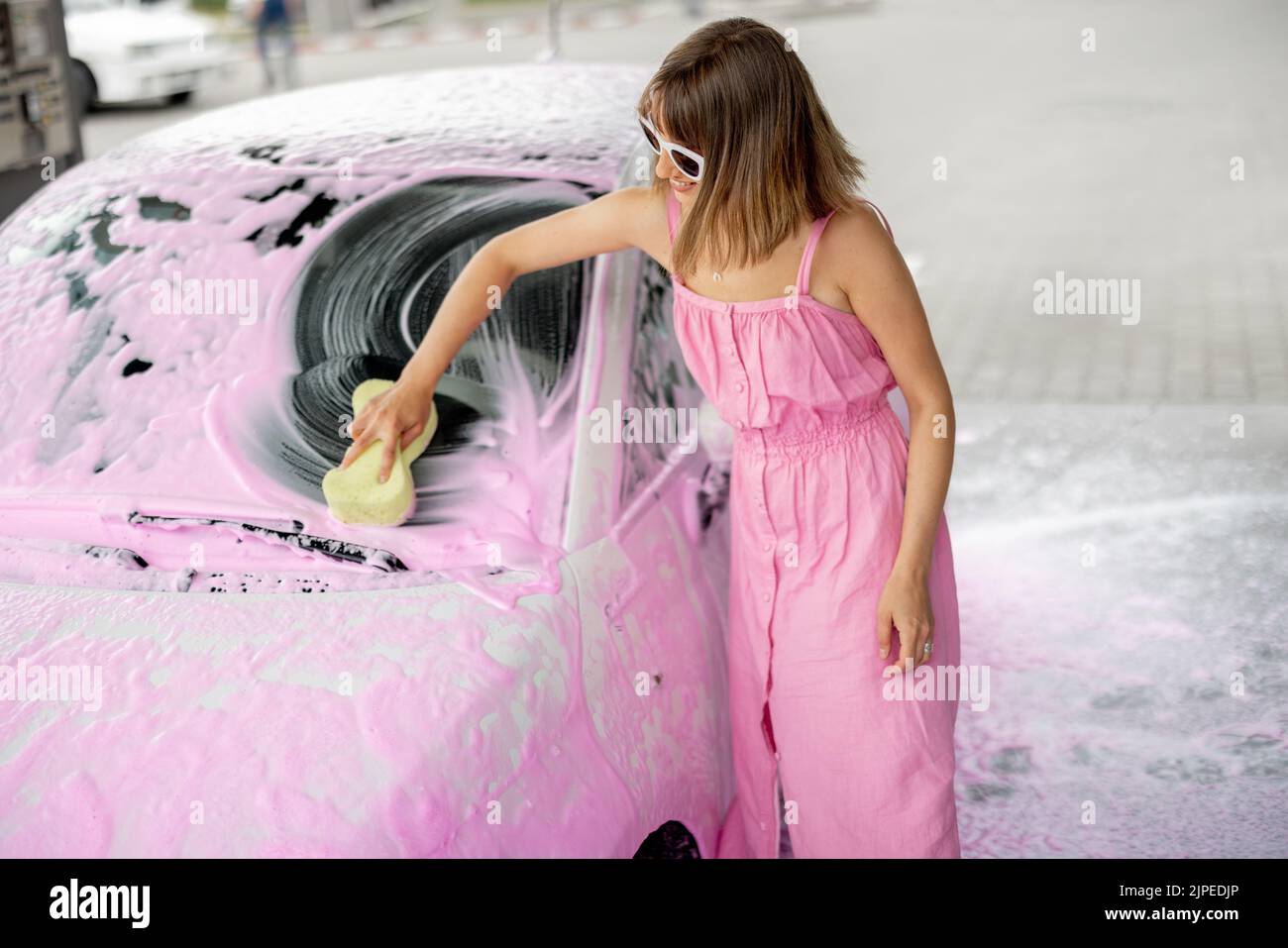 Young woman in pink dress wiping her tiny car covered in nano foam with a sponge at car wash. Concept of easy and beautiful self-service at car wash Stock Photo