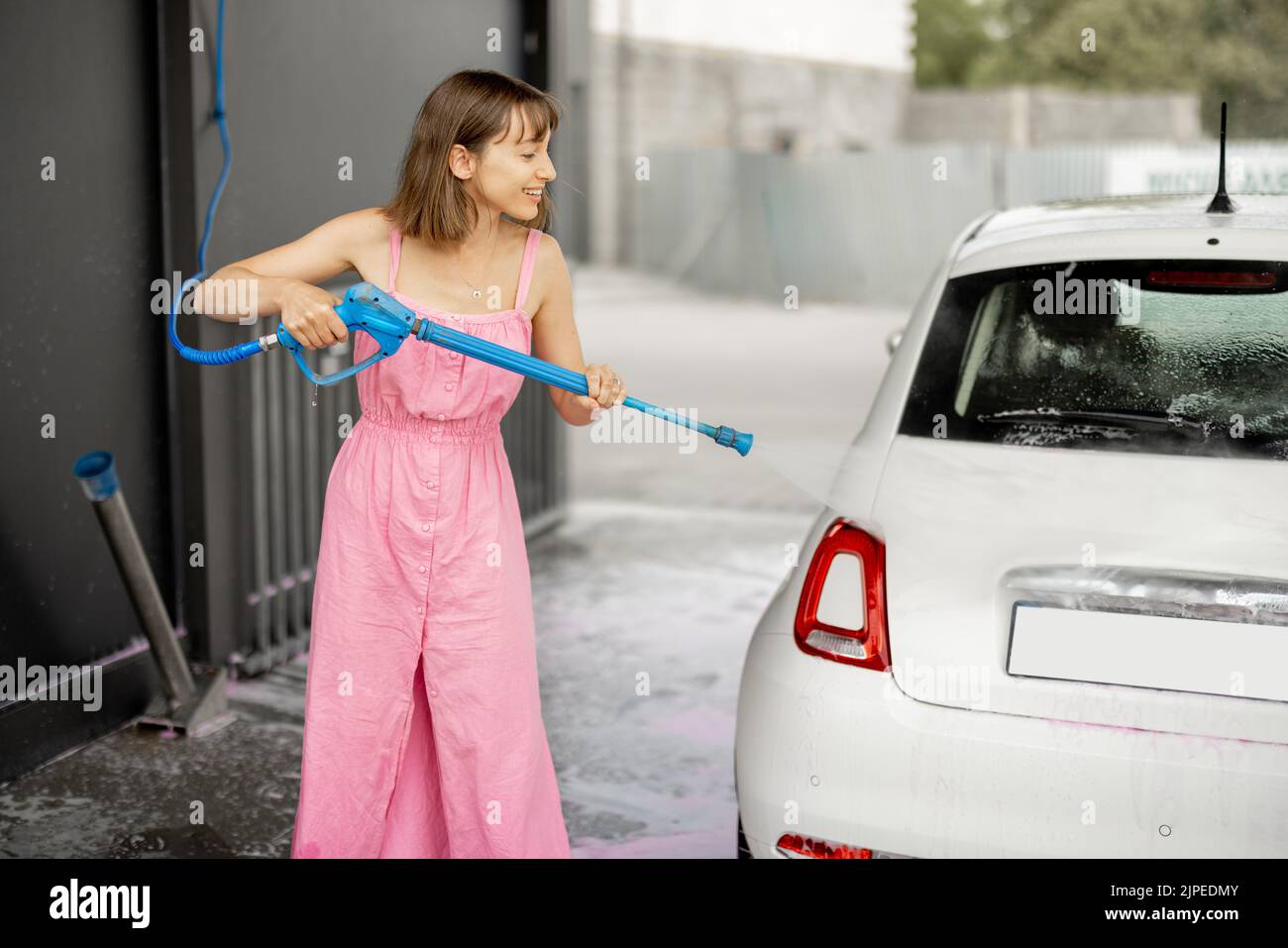 Young woman in pink dress washing her tiny vehicle with washing gun under the pressure at self-service car wash. Easy process of car care by female co Stock Photo