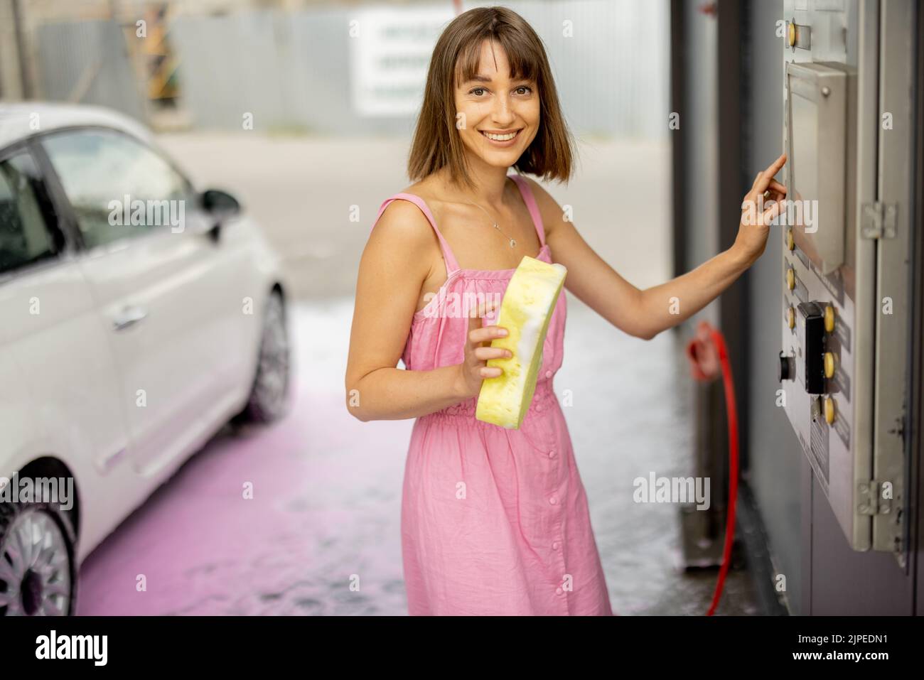 Young cheerful woman in pink dress pushing button on wash machine choosing the washing mode at car self-service Stock Photo