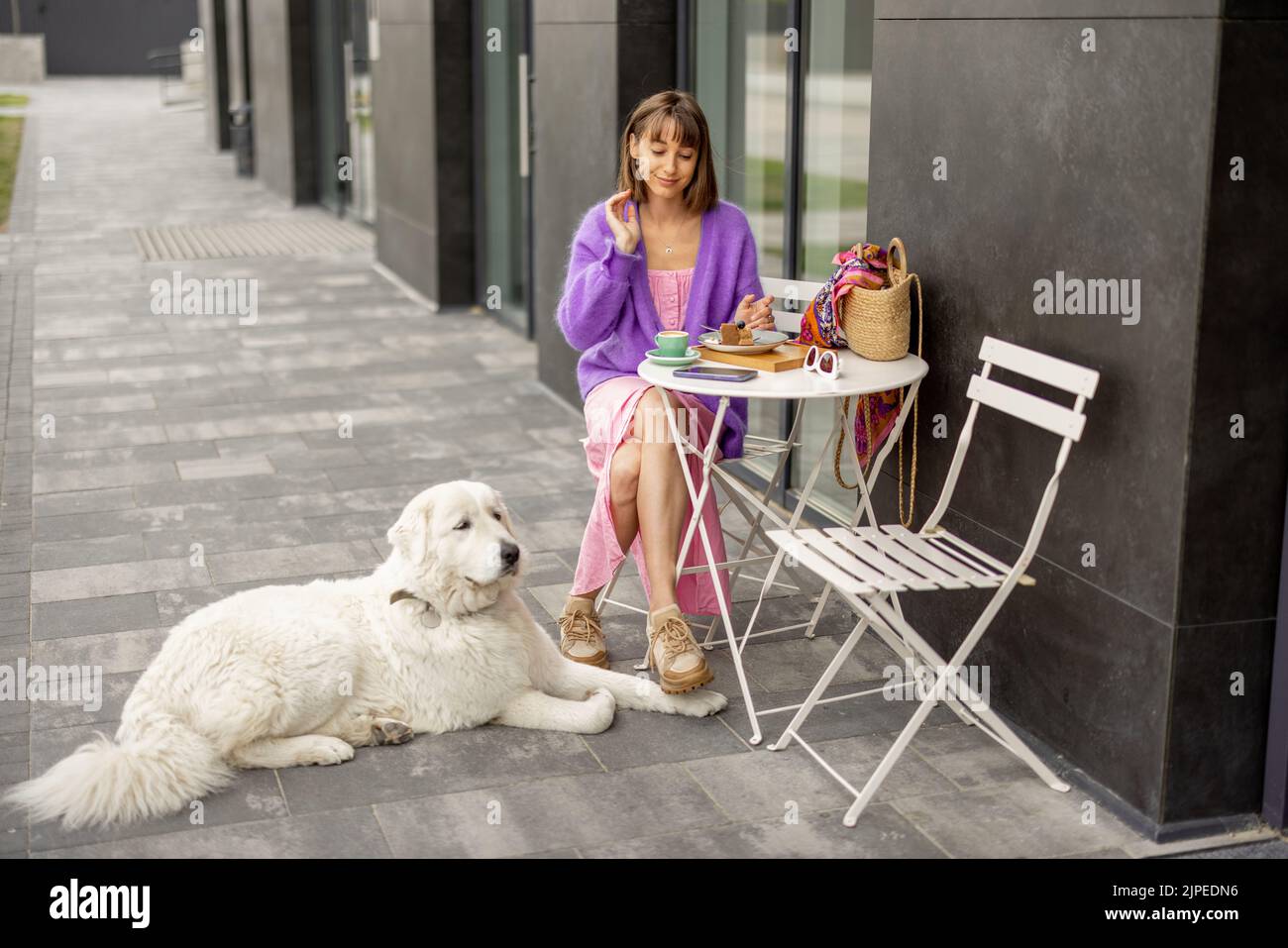 Young stylish woman sits with her cute adorable dog at cafe on a street. Leisure time with pet spending time at modern housing estate outdoors Stock Photo