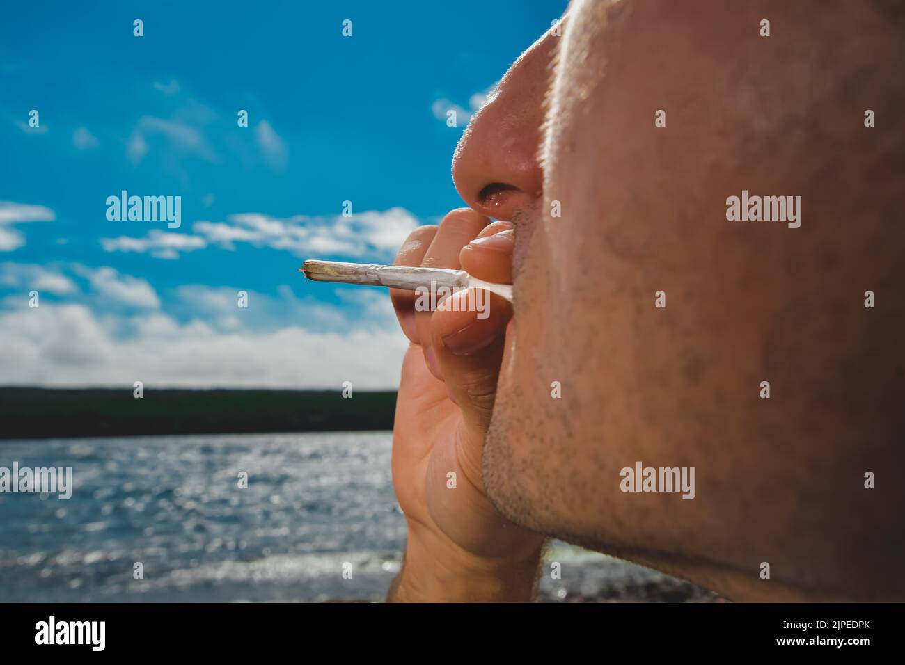 Detail of a man smoking a marijuana or weed joint relaxing on the shore of a lake. Stock Photo
