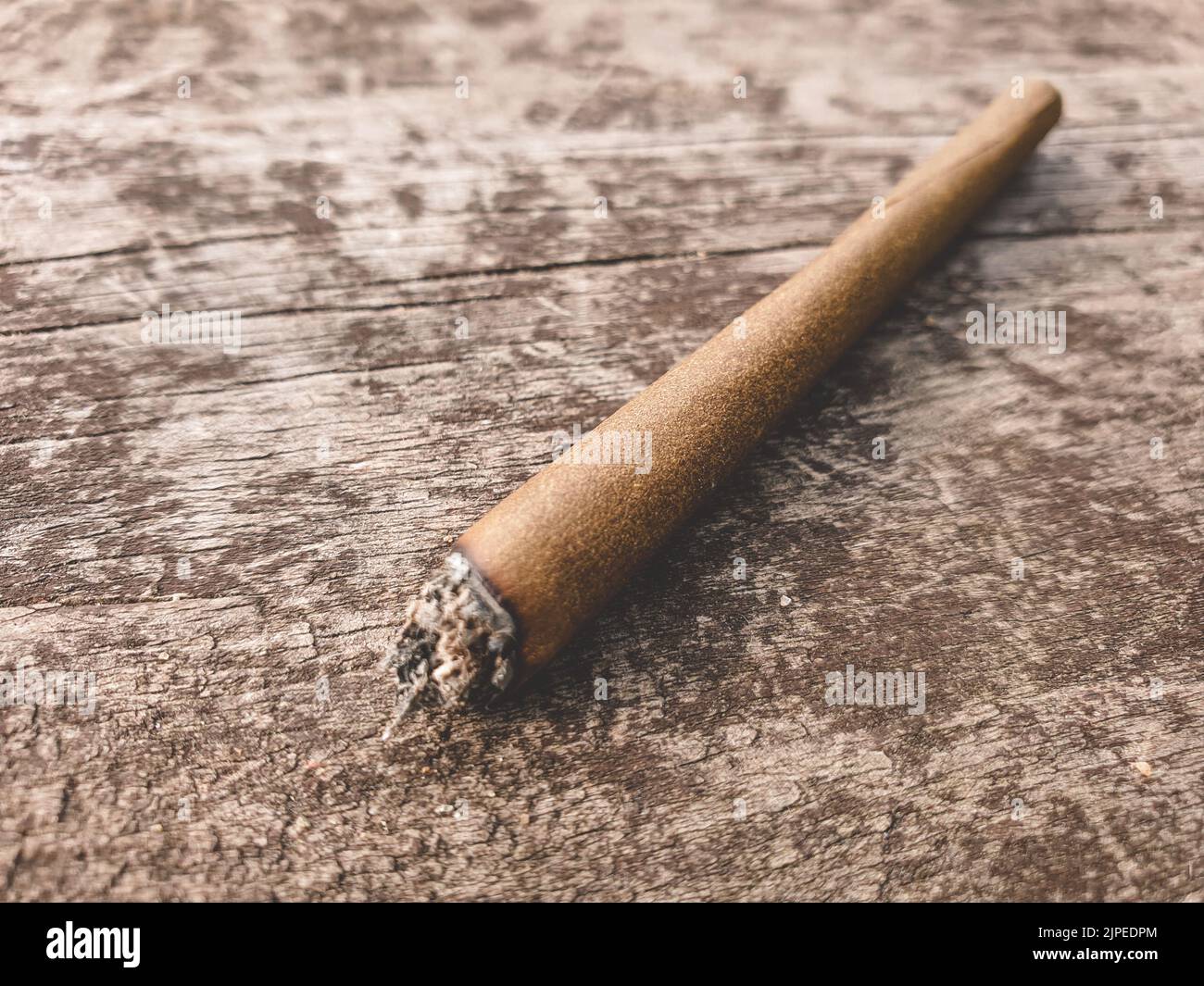 Detail of a lit marijuana or weed joint pictured on a brown wooden table outdoors. Stock Photo