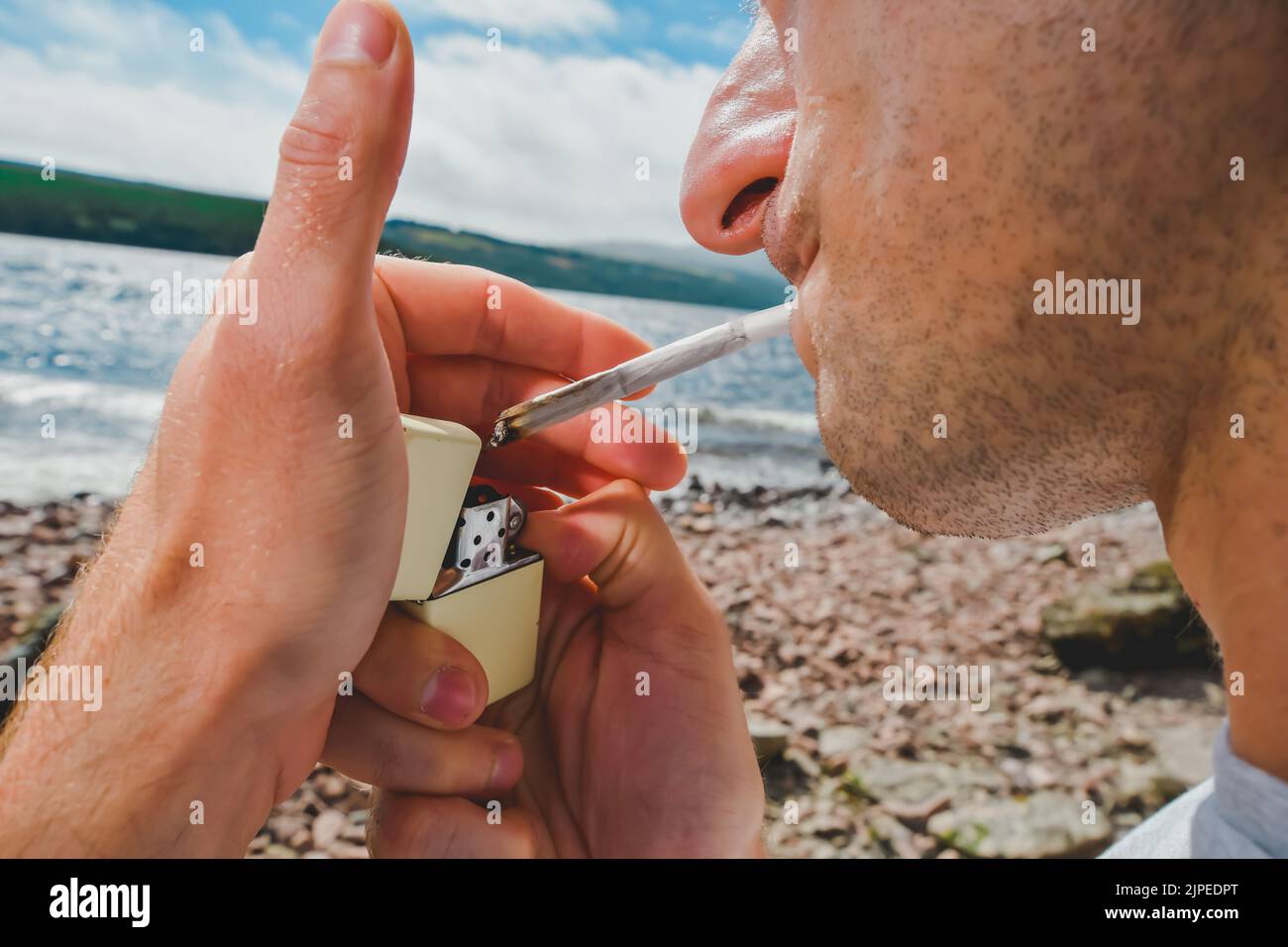 Detail of a man lighting up a marijuana or weed joint outdoors on the shore of a lake. Stock Photo