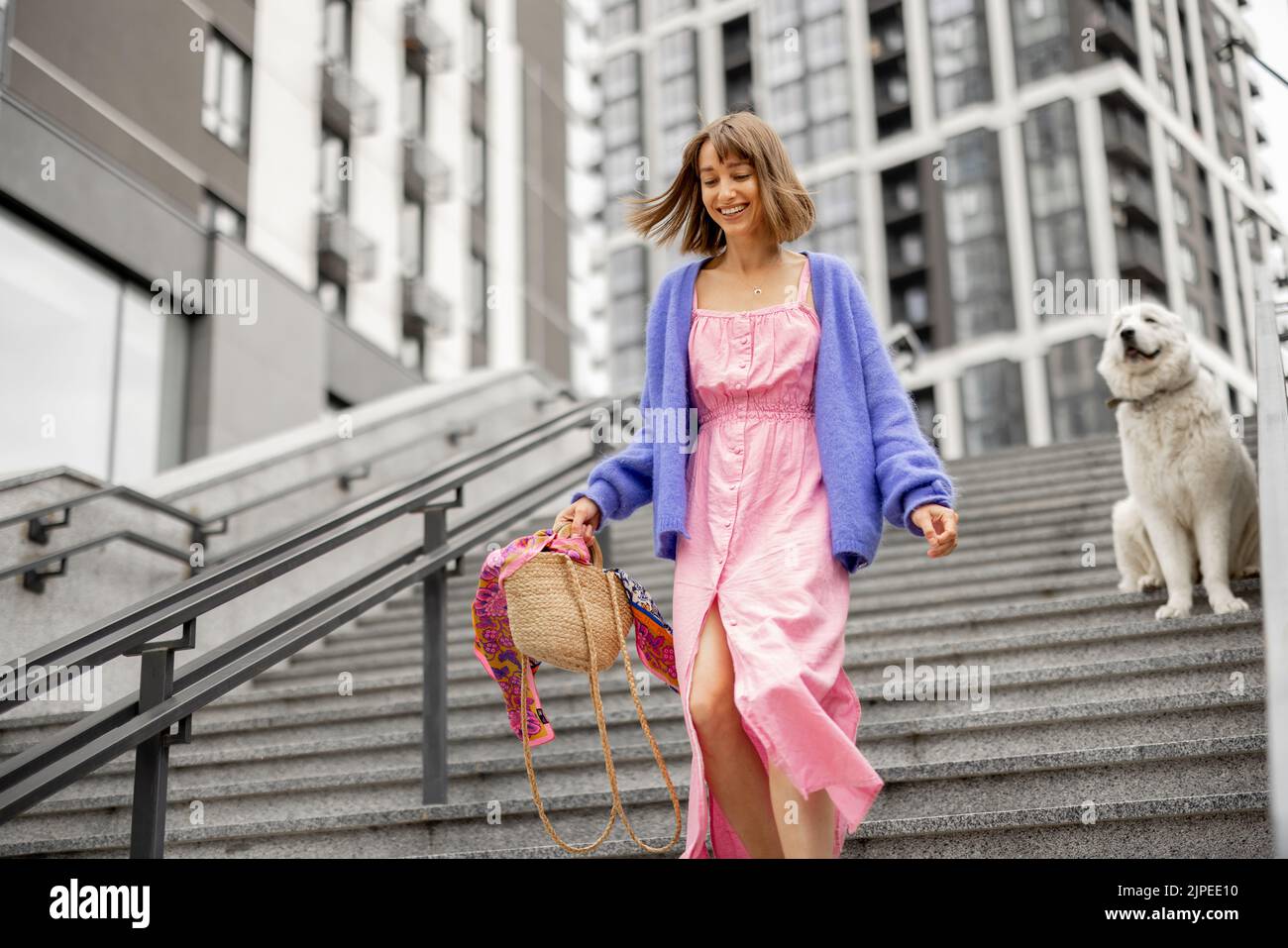 Young stylish woman dressed brightly runs down on staircase at modern residential area. Dog behind. Concept of style and carefree lifestyle Stock Photo