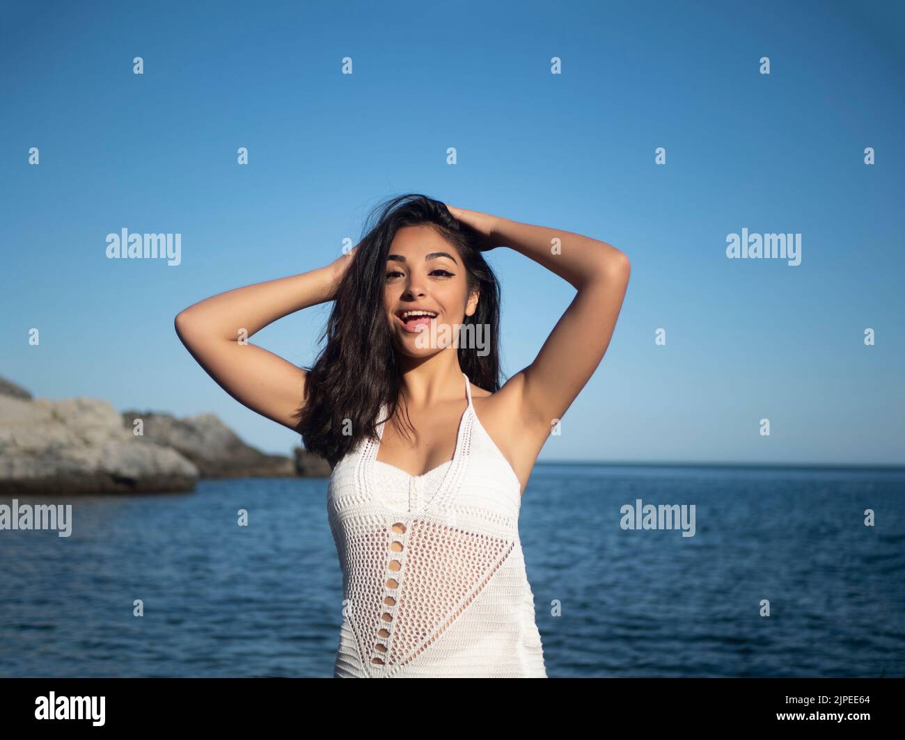 Self assured young Hispanic female tourist in white dress with long dark hair smiling and touching head while looking at camera on wavy seashore under Stock Photo