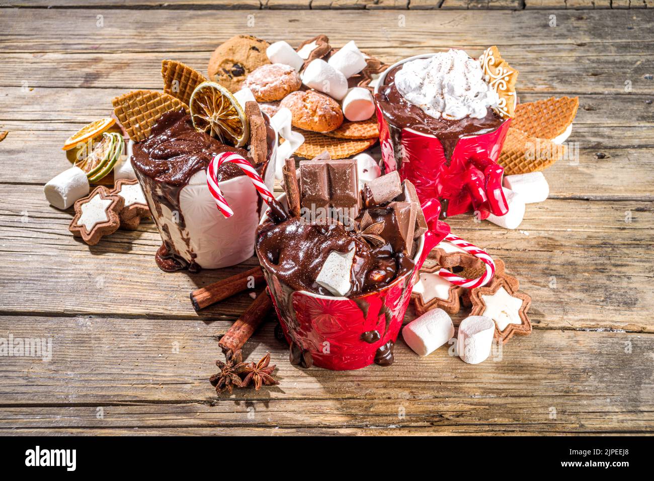 Christmas styled overloaded hot chocolate mugs with various toppings – chocolate slices, spices, sweets, cookies, candy and gingerbreads, wooden cozy Stock Photo