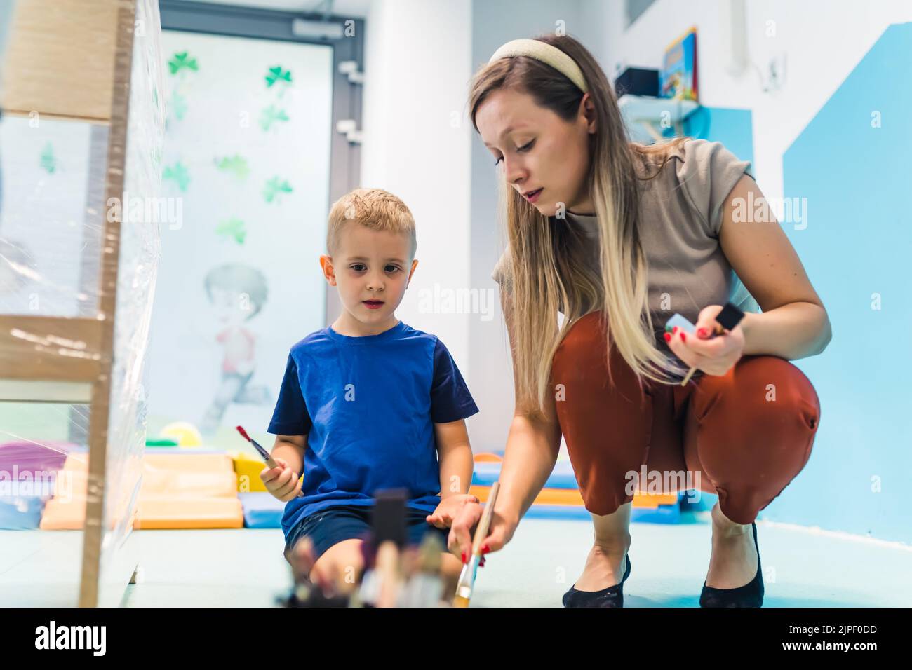 Nursery school. Caucasian toddler boy and his teacher painting on a cling film wrapped around the wooden shelving stand by using brushes and nontoxic paints . Fun game activity for kids sensory skills, creativity, spatial attention and visual perceptual development . High quality photo Stock Photo