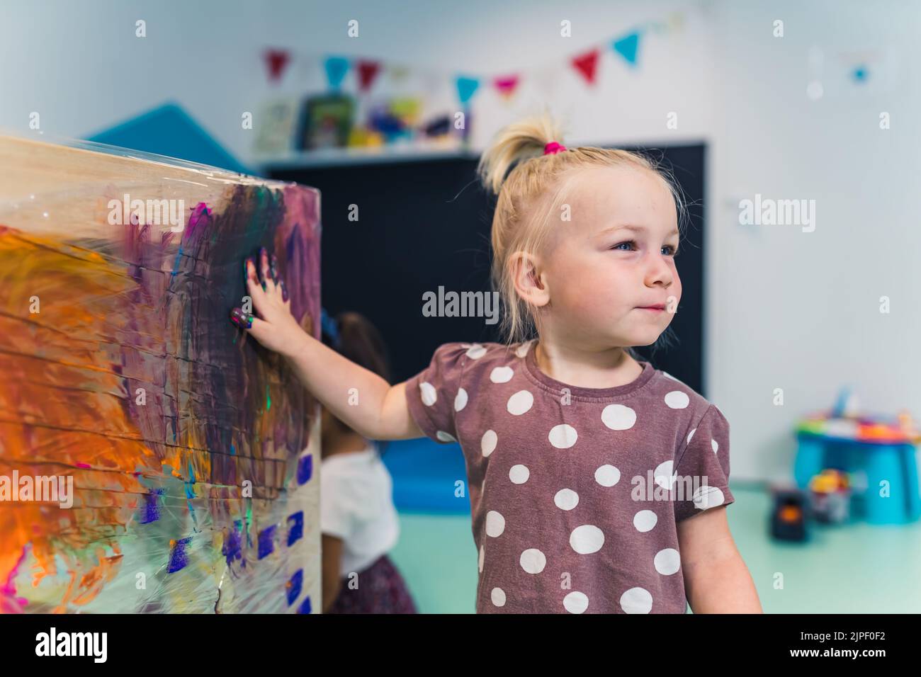 Nursery school. Happy caucasian blonde toddler girl finger painting on a cling film wrapped around the shelving stand as an easel. Fun art activity for kids sensory skills, creativity and imagination development . High quality photo Stock Photo