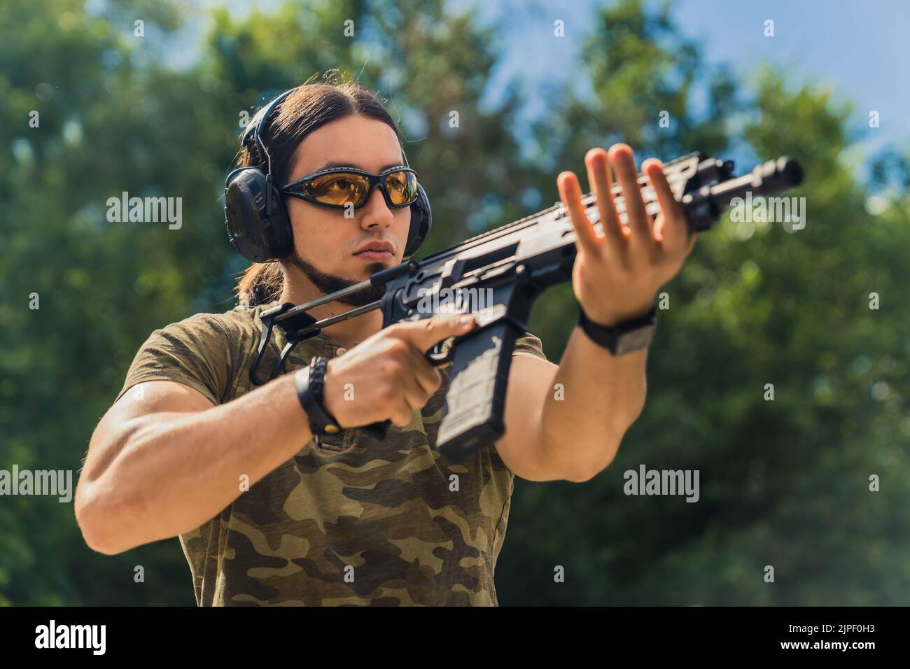 Bearded man in safety goggles headphones and camo t-shirt practicing using submachine gun. Outdoor firing range. Horizontal shot. High quality photo Stock Photo