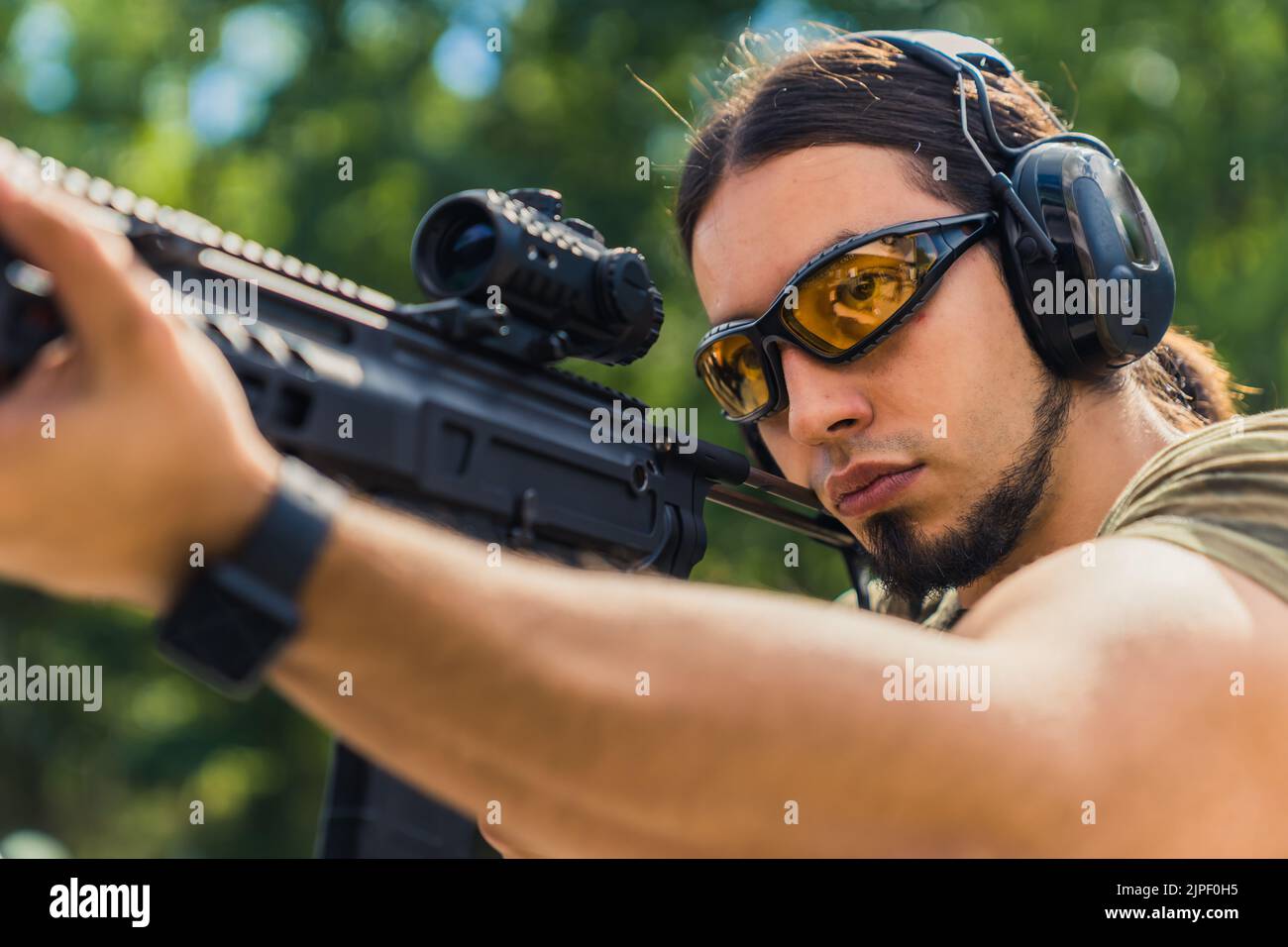 White man with beard in protective goggles and headphones aiming submachine gun. Outdoor firing range. Horizontal shot. High quality photo Stock Photo