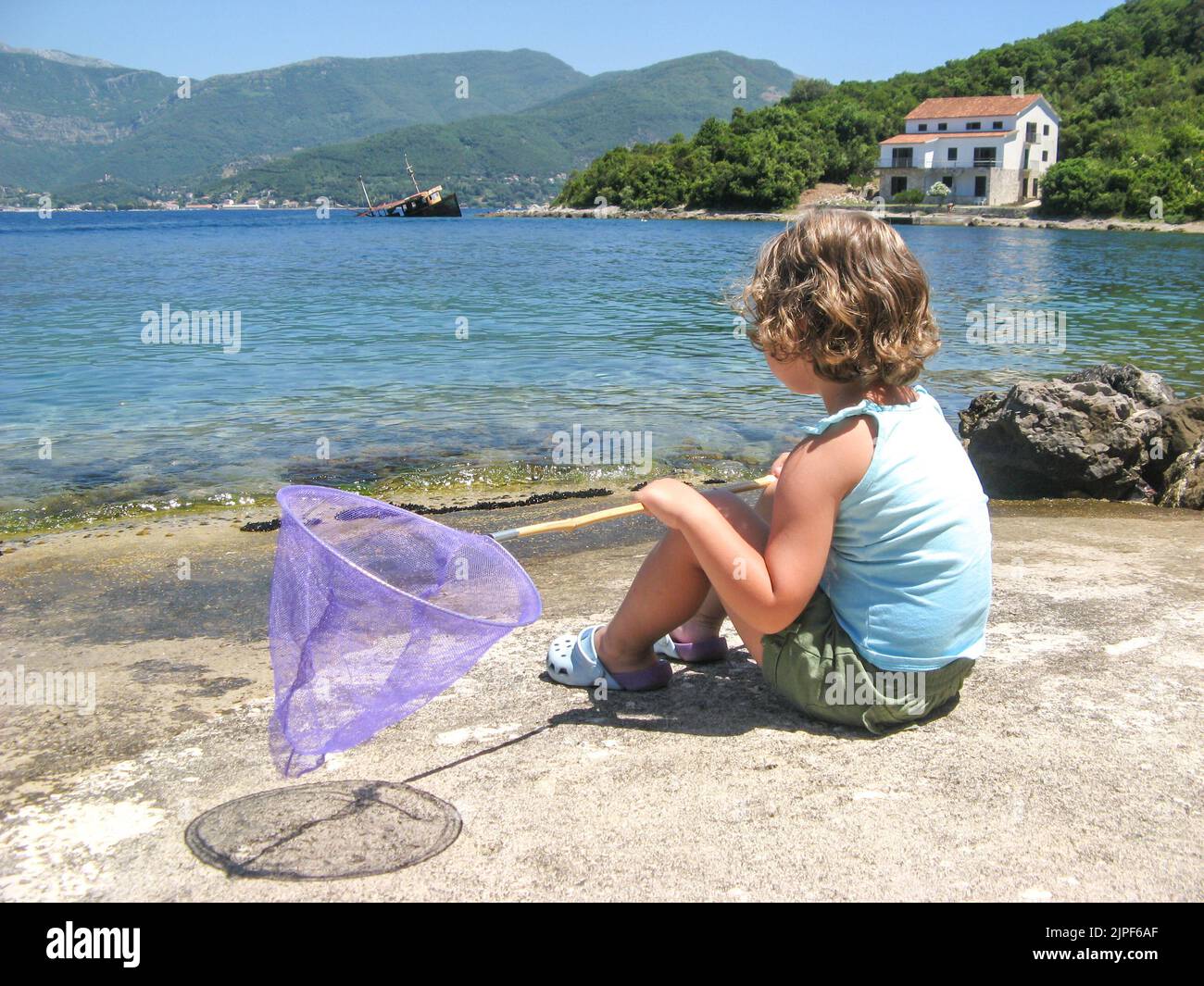 Cute curly toddler girl with butterfly net for catching fish sitting on the seashore, lookig at sea. Stock Photo