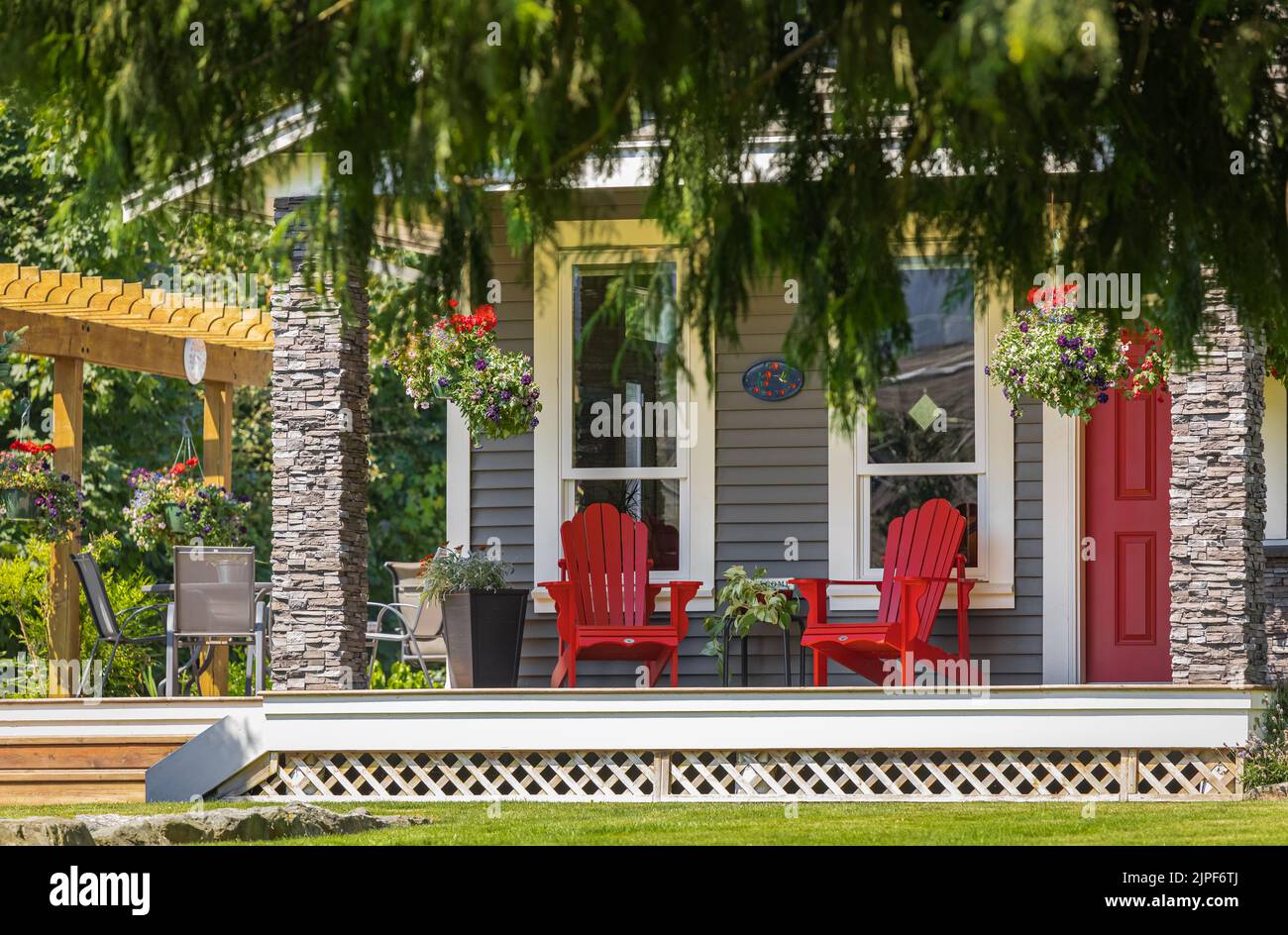 View of a large front porch with furniture and potted plants. Rustic, country style front porch seating with red Adirondack chairs and fresh flowers. Stock Photo