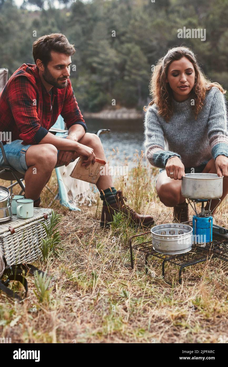 Young couple camping by the lake. Young woman preparing food on camping stove with her boyfriend sitting by. Stock Photo