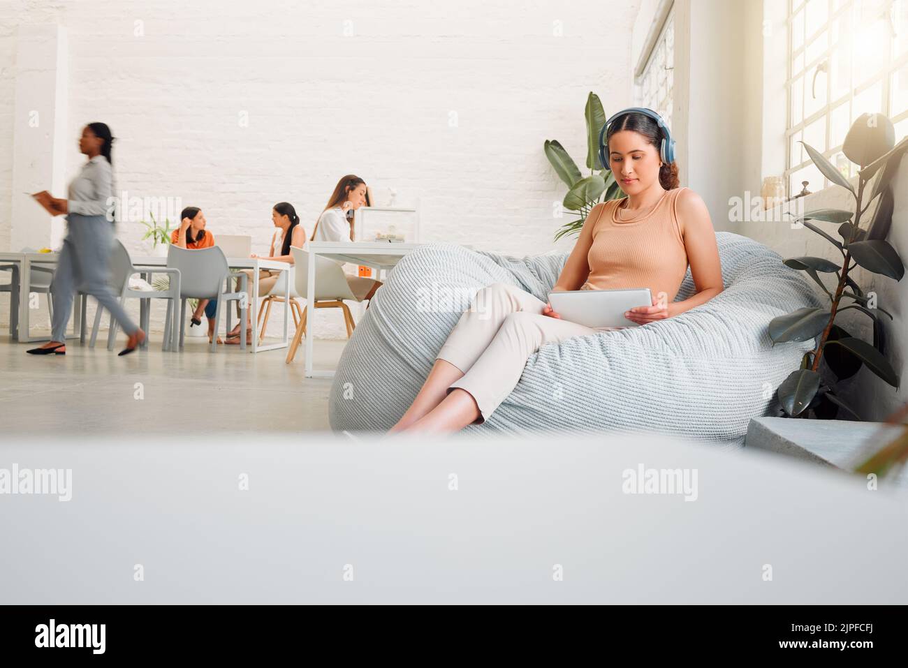 Creative intern browsing online with a tablet, listening to music on wireless headphones. Female on a break sitting on bean bags in a casual office Stock Photo