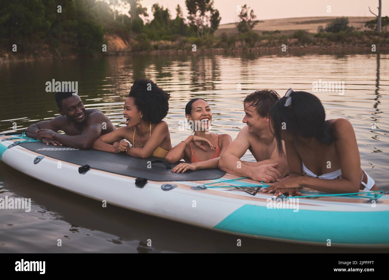 Friends, vacation and having fun while leaning on a paddle board and talking in a lake. Happy and diverse people laughing while enjoying the water and Stock Photo