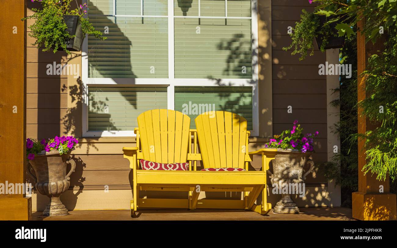 View of a large front porch with furniture and potted plants. Rustic, country style front porch seating with yellow Adirondack chairs and fresh flower Stock Photo