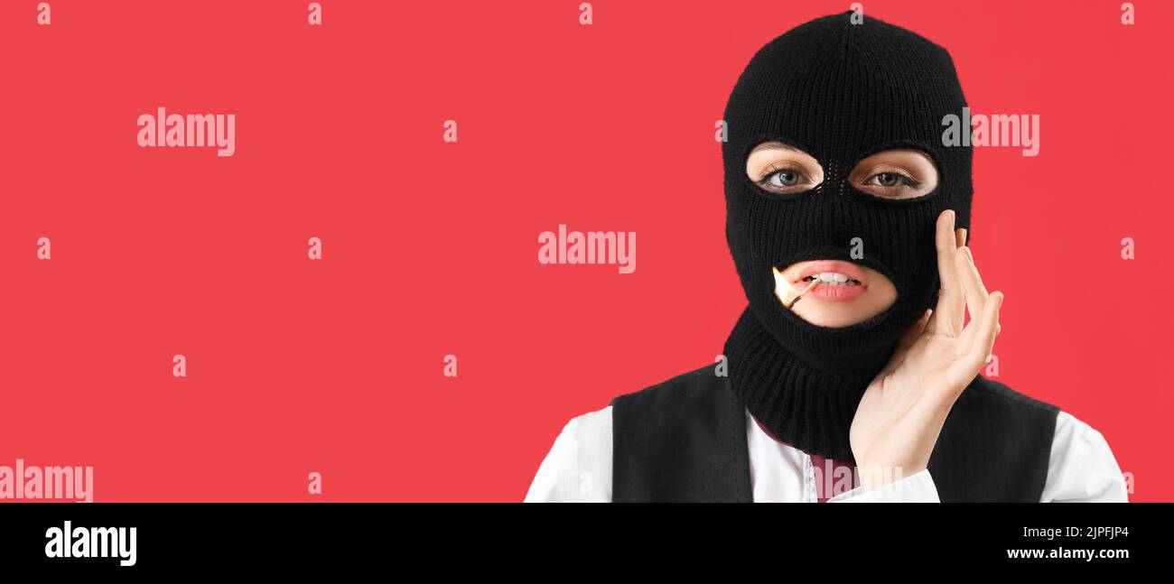 Portrait of young woman in balaclava and with burning match in mouth on red background with space for text Stock Photo