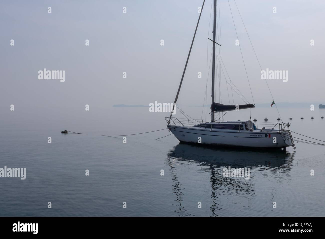 Landscapes of Lake Garda on a foggy day, with boats, birds, cypress trees, reflections on the calm white water, calm after the storm, metaphysical Stock Photo