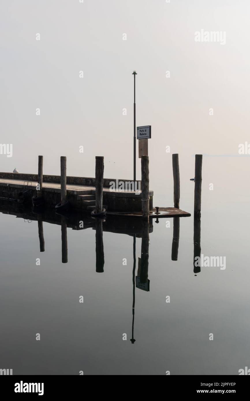 Landscapes of Lake Garda on a foggy day, with boats, birds, cypress trees, reflections on the calm white water, calm after the storm, metaphysical Stock Photo