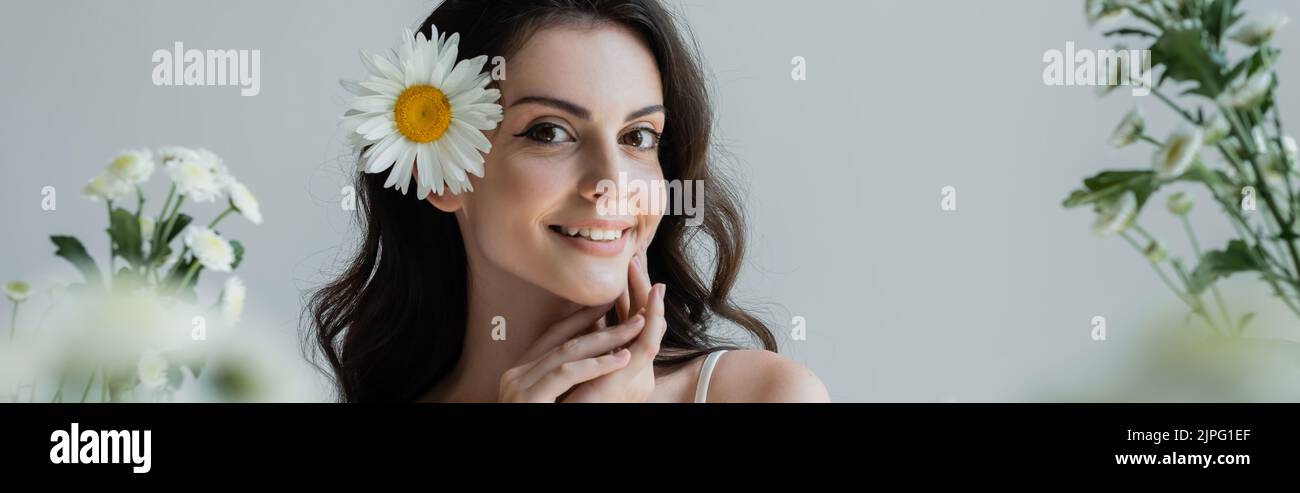 Young woman with makeups and chamomile in hair smiling at camera near blurred flowers isolated on grey, banner Stock Photo