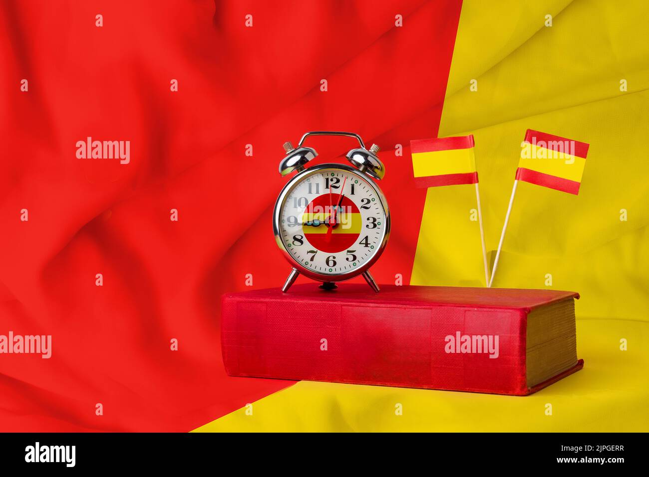 Time to learn Spanish. A clock on a book with two Spanish flags on a red and yellow background. Studying one of the most spoken languages in the world Stock Photo
