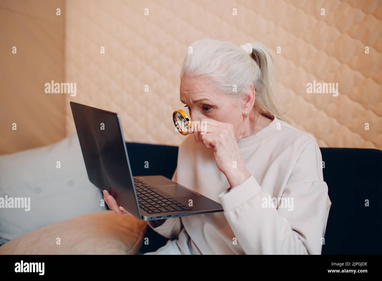 Elderly woman grandmother learns to work at home on laptop computer and internet with magnifier. Stock Photo