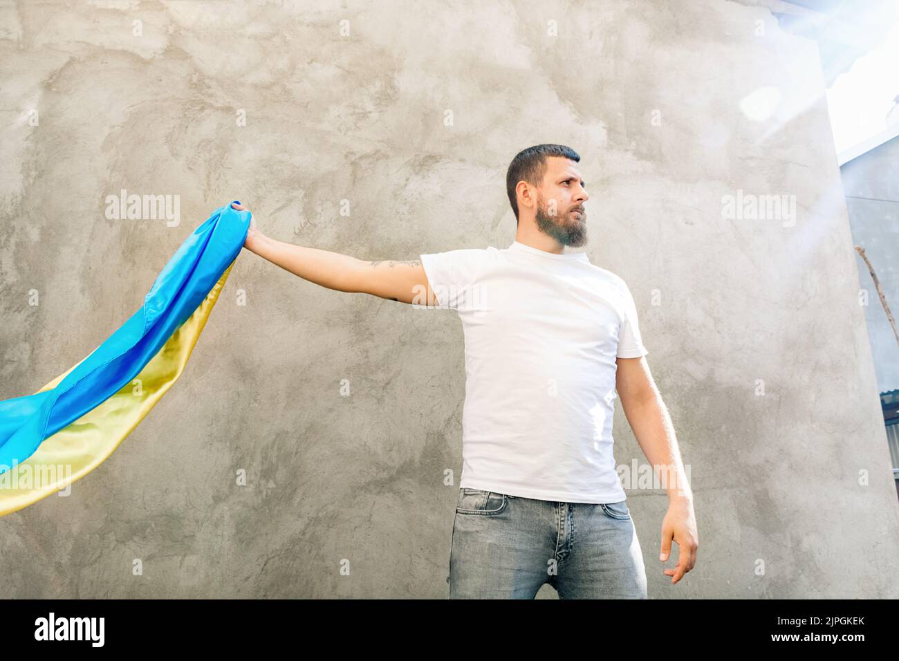 Man holds in his hand the State Blue and Yellow Flag of Ukraine. Guy in white T-shirt and jeans. Sunny day. Constitution and Independence Day of Ukraine. Bottom view of photo. Soft Selective focus Stock Photo