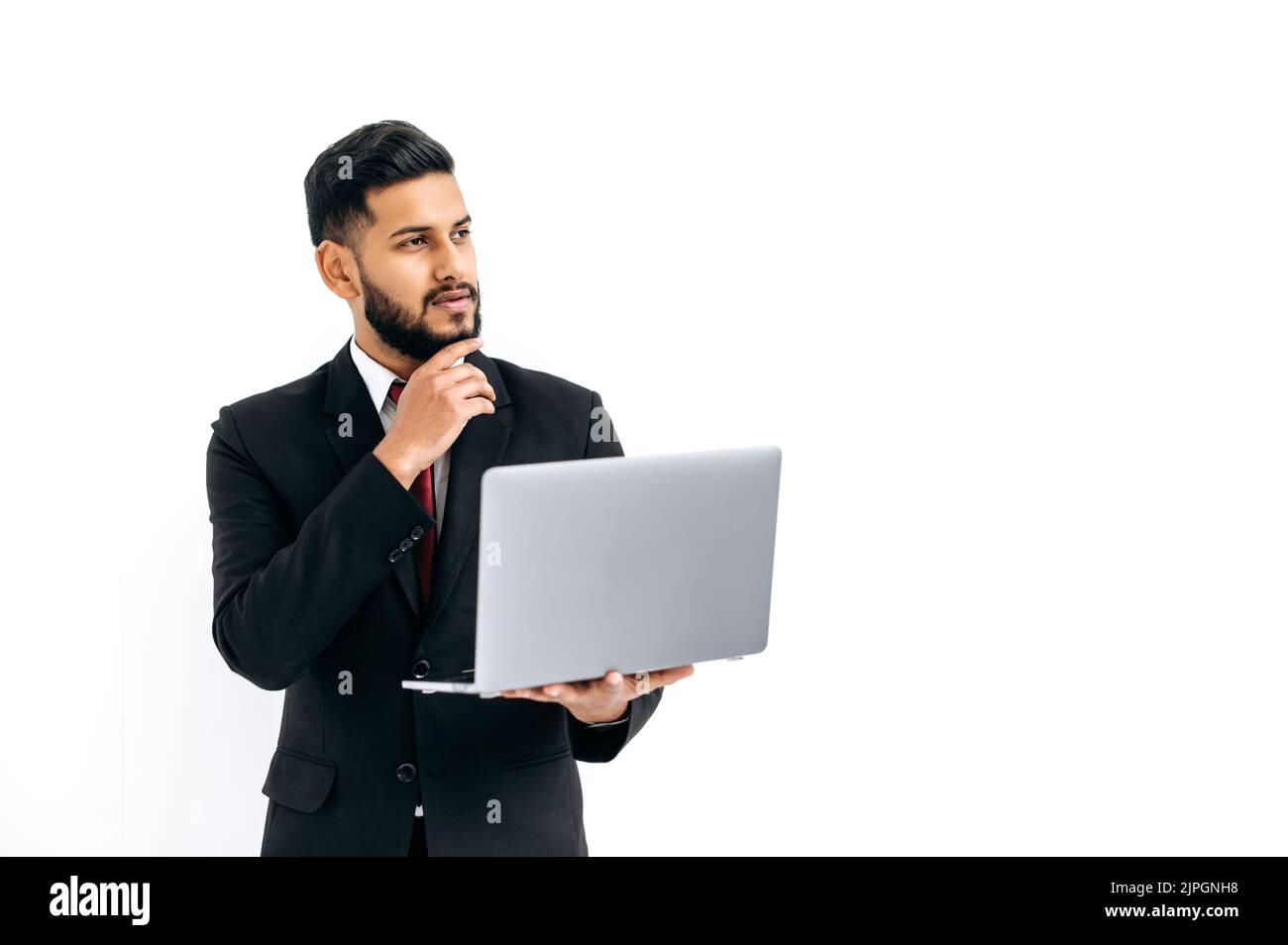 Puzzled thoughtful elegant influential arabian or indian man in formal suit, stand on isolated white background with laptop, pensively looking away, thinking, dreaming.Copy-space for your presentation Stock Photo