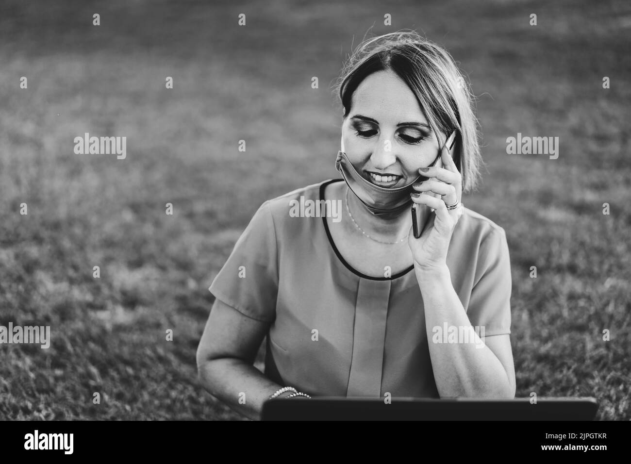 Woman 40 years old working outdoors with laptop during coronavirus outbreak - Smiling entrepreneur sitting on lawn with personal computer while callin Stock Photo