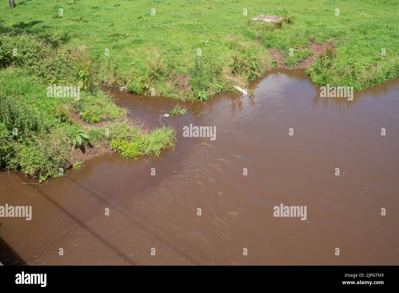 A tributary polluted with farm ellfluent discharges dark brown water into the already silt and effluent laden Gwendraeth Fach river Stock Photo