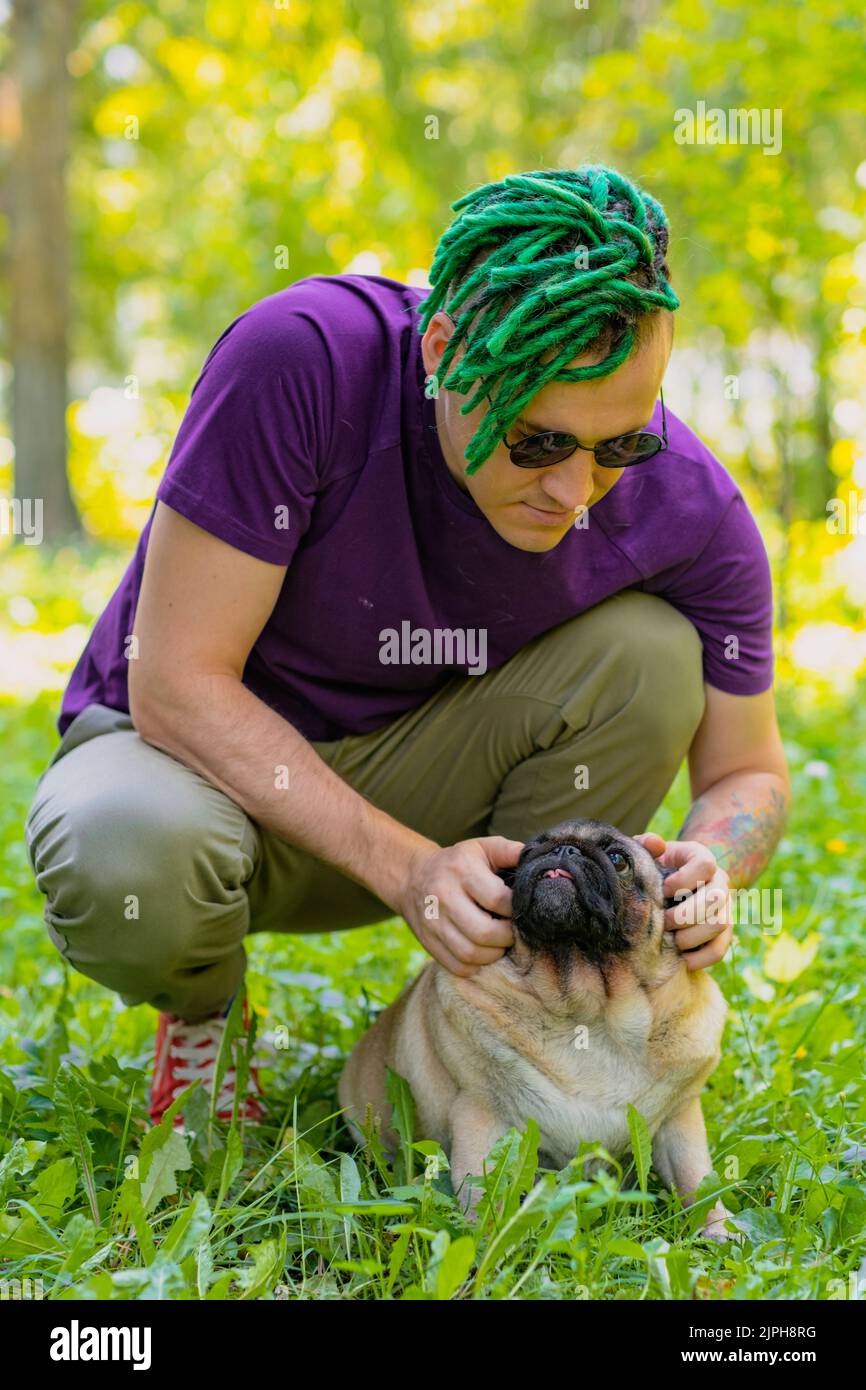 A hipster with green dreadlocks and black round glasses is sitting on a green lawn and stroking a cute pug on a sunny summer day. A man walks a pug Stock Photo